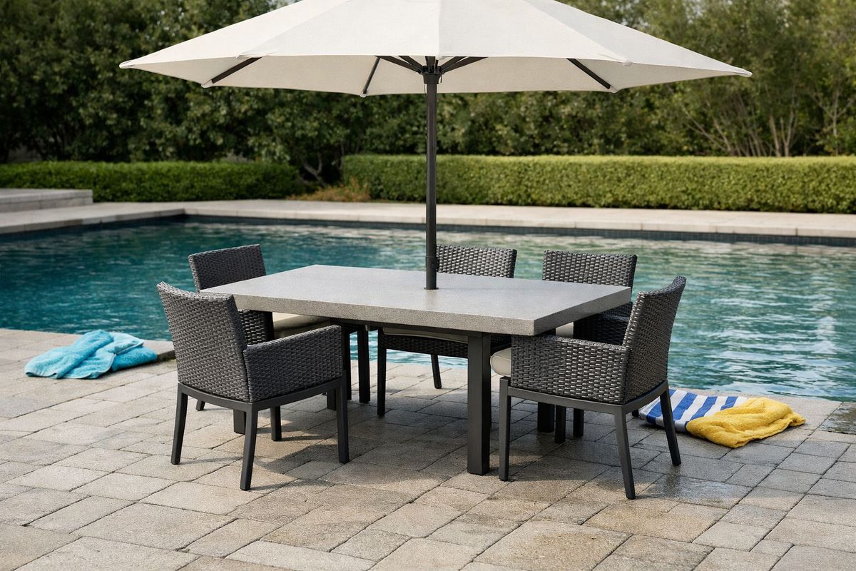 A poolside dining area with a rectangular table made of sealed stone or composite material, four weather-resistant chairs, a large umbrella casting shade over the table, and slip-resistant pavers underfoot, set beside a swimming pool with wet deck edges and a few scattered towels nearby.