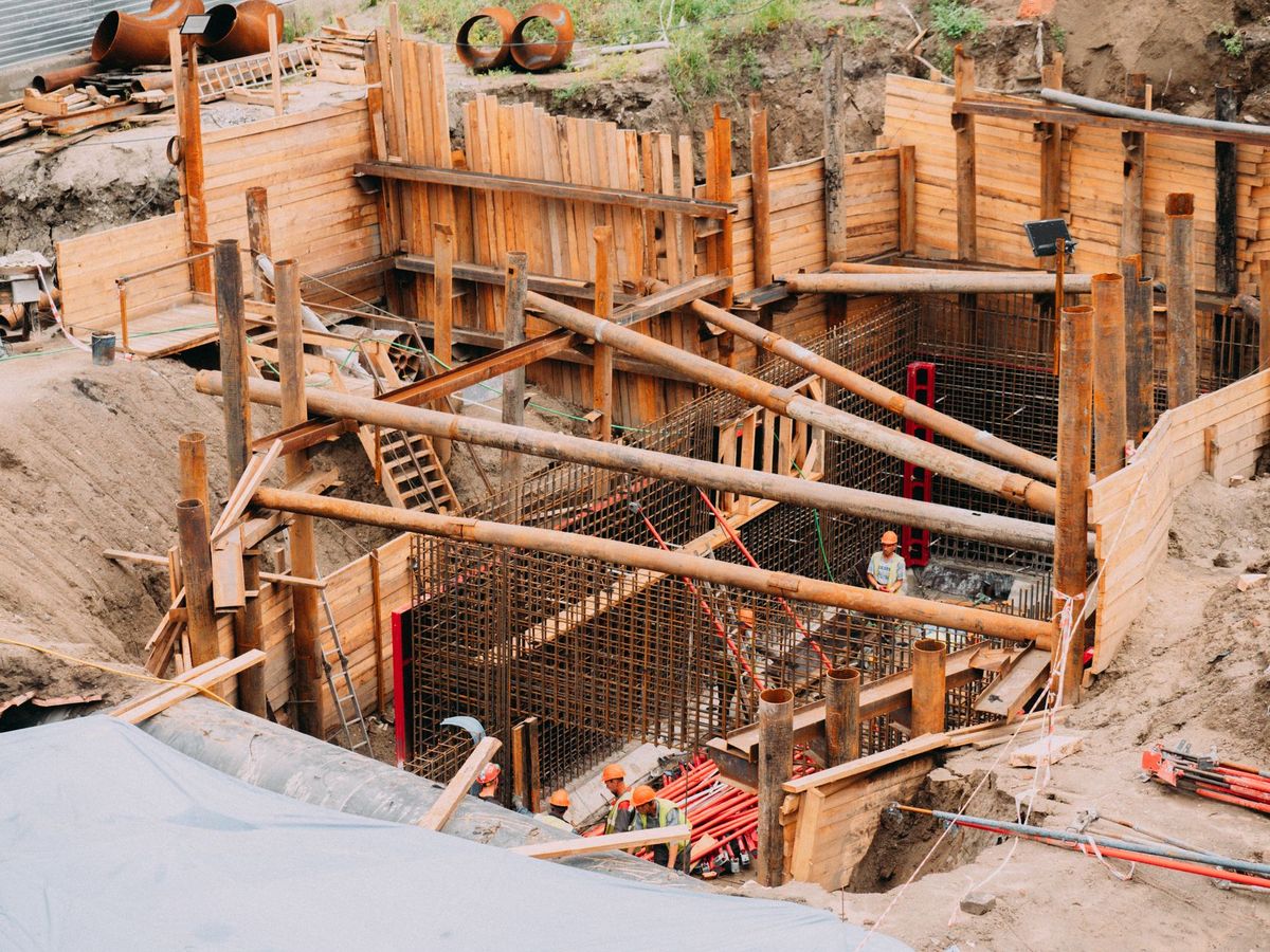 A backyard pool construction site in progress with an excavated hole, steel reinforcement grid, PVC plumbing pipes, electrical conduit, stacked coping stones, and workers’ tools arranged around a partially built pool shell, with a house and patio visible in the background