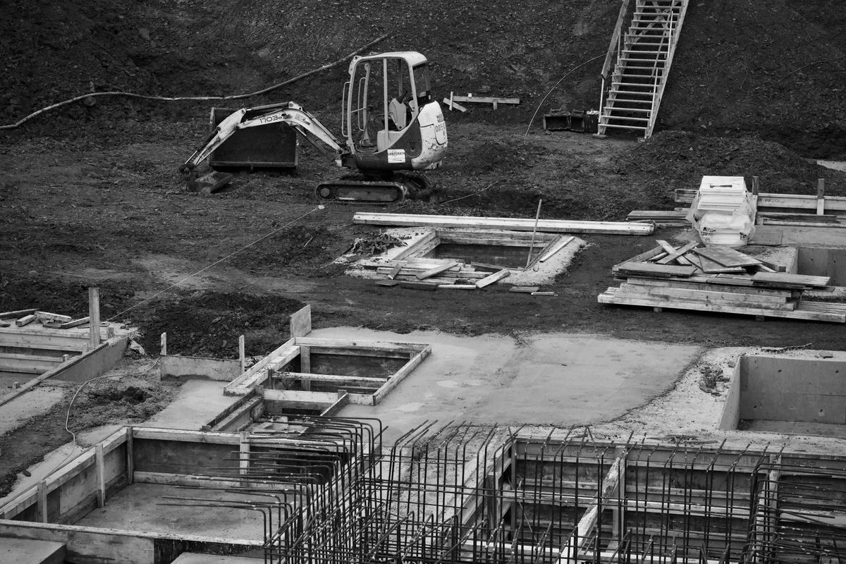 A backyard pool construction site split between two stages: on one side, workers are spraying a gray gunite shell over steel rebar in an excavated hole with piles of dirt nearby; on the other side, a glossy blue fiberglass pool shell is suspended by a crane and being lowered into a dug-out yard beside stacked decking materials