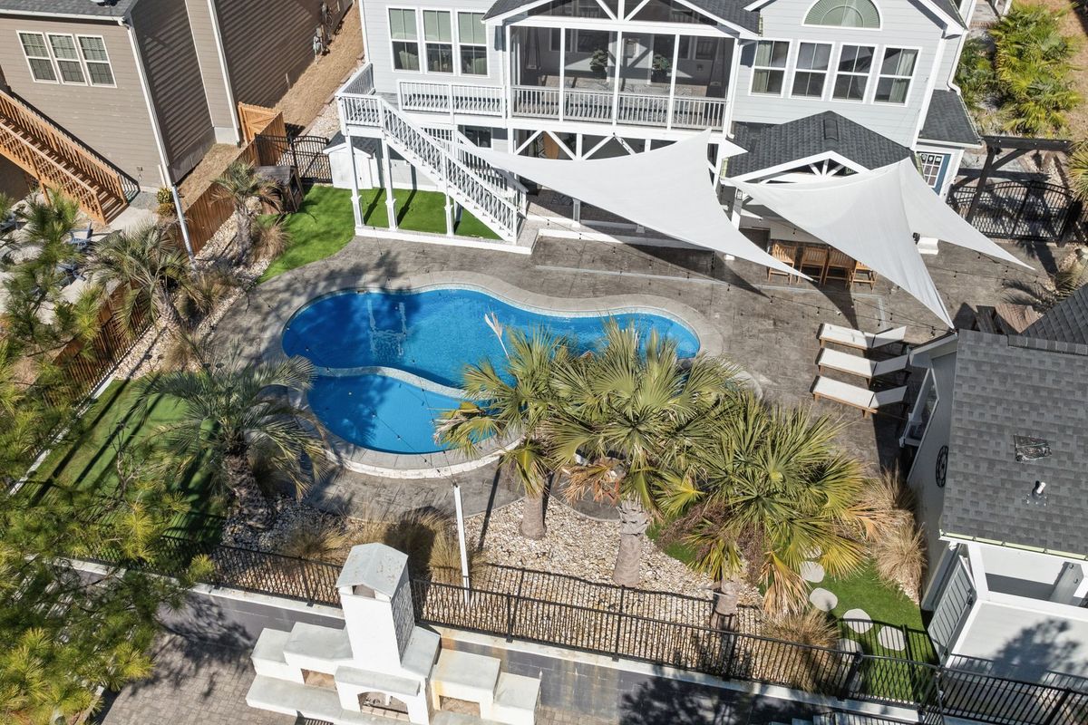 A Florida backyard pool with light-colored paver decking, a shaded covered patio area, a screened enclosure over part of the pool, rainwater draining toward a channel away from the house, and tropical landscaping around the edges under bright midday sun