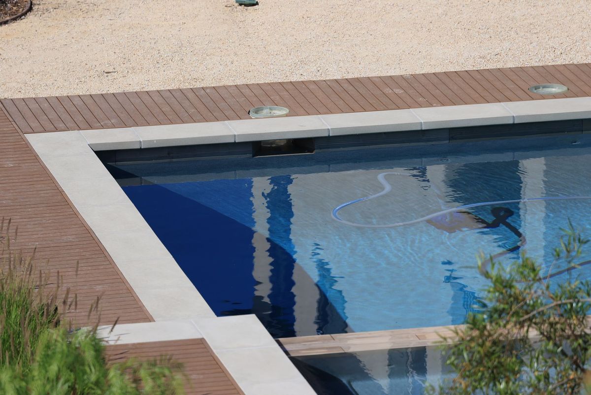 A backyard swimming pool with a skimmer net resting beside the deck, an open pool pump basket, a pool brush leaning against the coping, and a pool vacuum hose stretched across the water, showing a basic weekly cleaning setup around the pool.