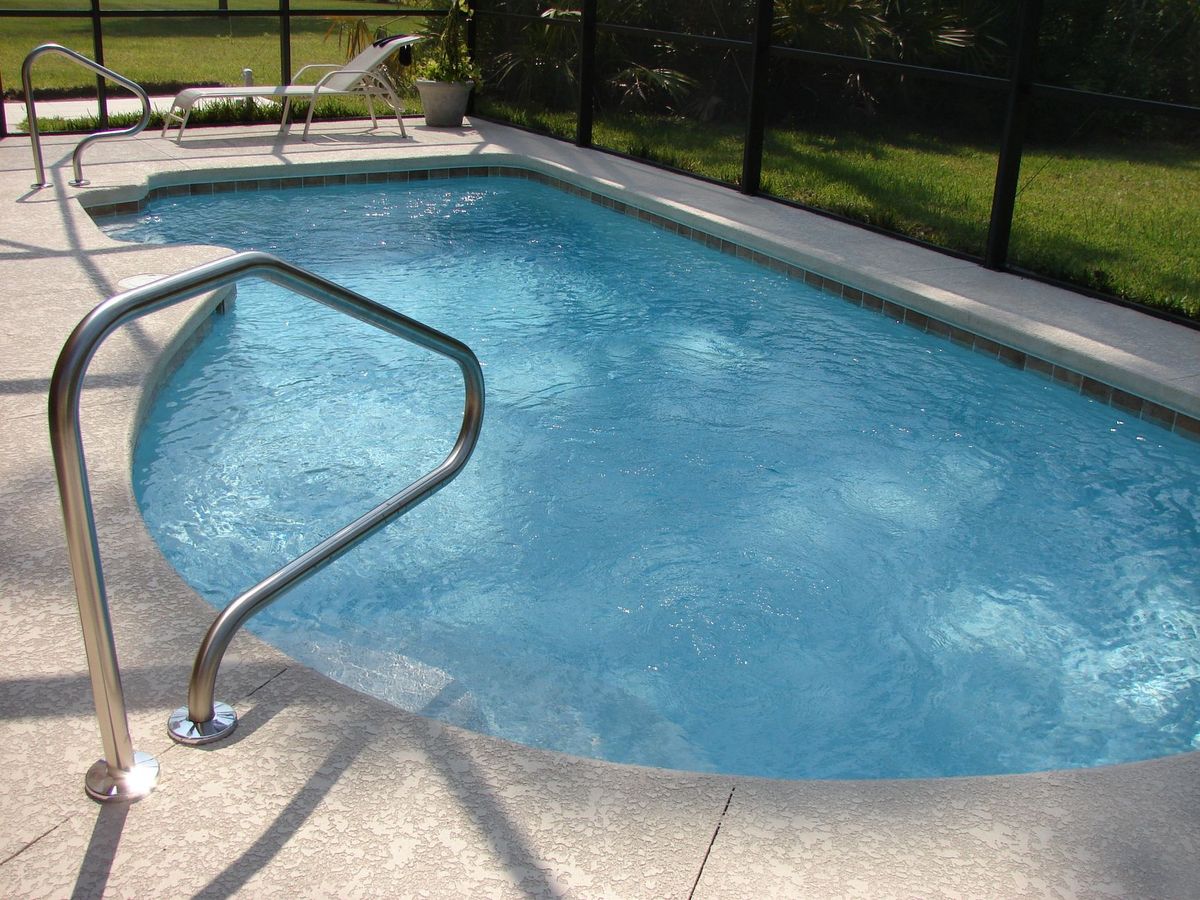 A clean rectangular backyard pool with visible skimmers, a pool filter and pump equipment pad beside the house, non-slip decking, a handrail at the entry steps, and trimmed landscaping kept back from the water to reduce debris