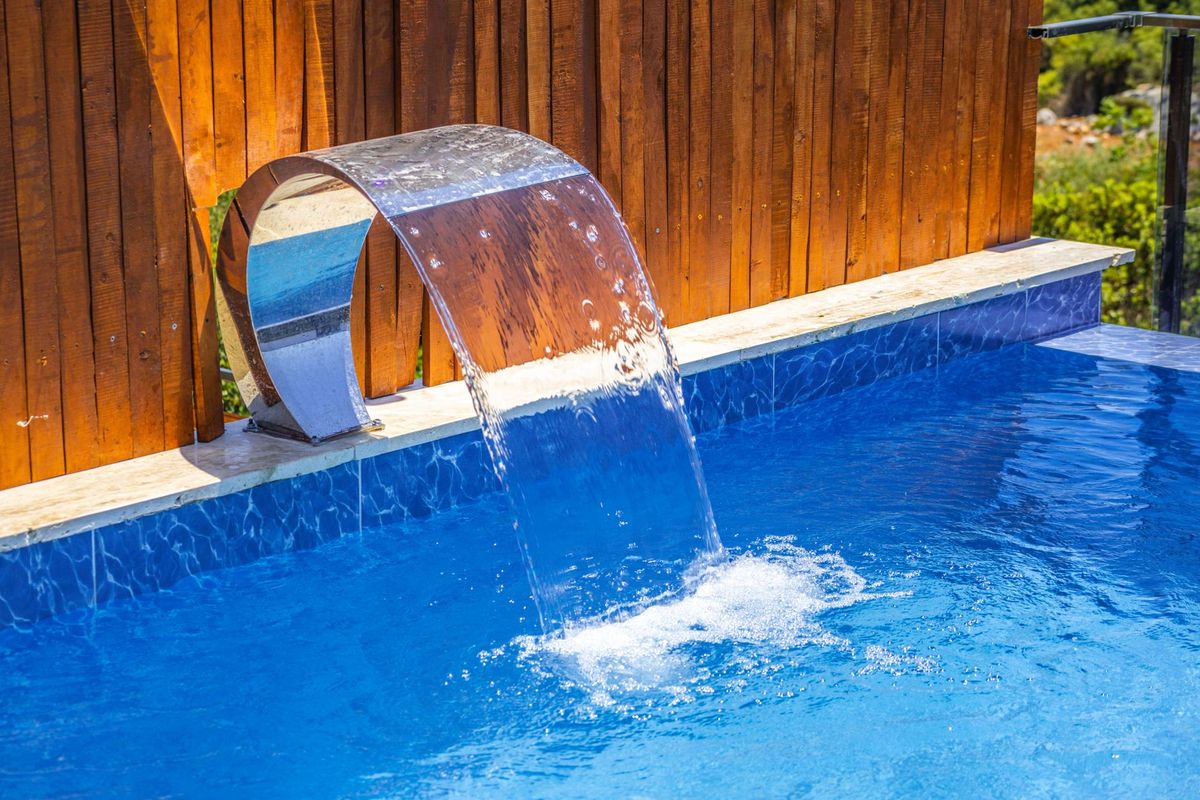 A custom backyard pool with an attached spa, a shallow sun shelf with bubblers, LED lights glowing underwater at dusk, a raised stone water feature spilling into the pool, and wide paver decking leading toward a screened outdoor living area