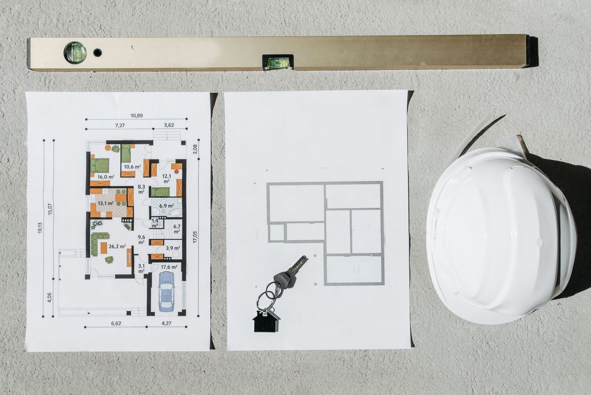 A large flat site plan sheet spread across a worktable beside measuring tools and a rolled-up property survey, showing a house footprint, driveway, property boundaries, an outlined pool location, setback measurements, a fence layout, and utility lines drawn across the yard area.