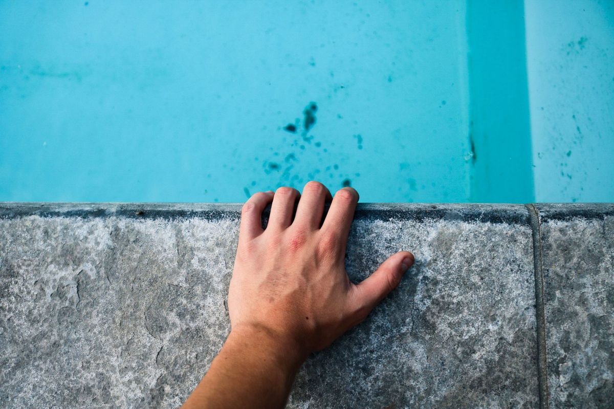 A close-up outdoor scene of three different pool water-edge surfaces: a textured concrete pool step with a hand brushing algae from the surface, a glossy smooth fiberglass ledge with clear water reflecting sunlight, and a vinyl liner pool edge with a soft blue liner and a visible seam, all beside a sunlit deck showing how the different materials feel and look in use