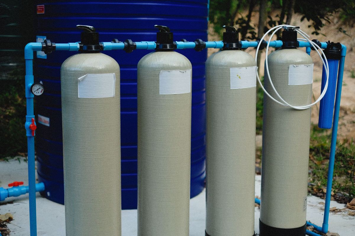 A close view of pool equipment with a pump, filter tank, pressure gauge, return pipes, and a clear plastic pump lid showing water and a few air bubbles, with a garden hose and cleaning tools nearby in a utility area beside the pool.